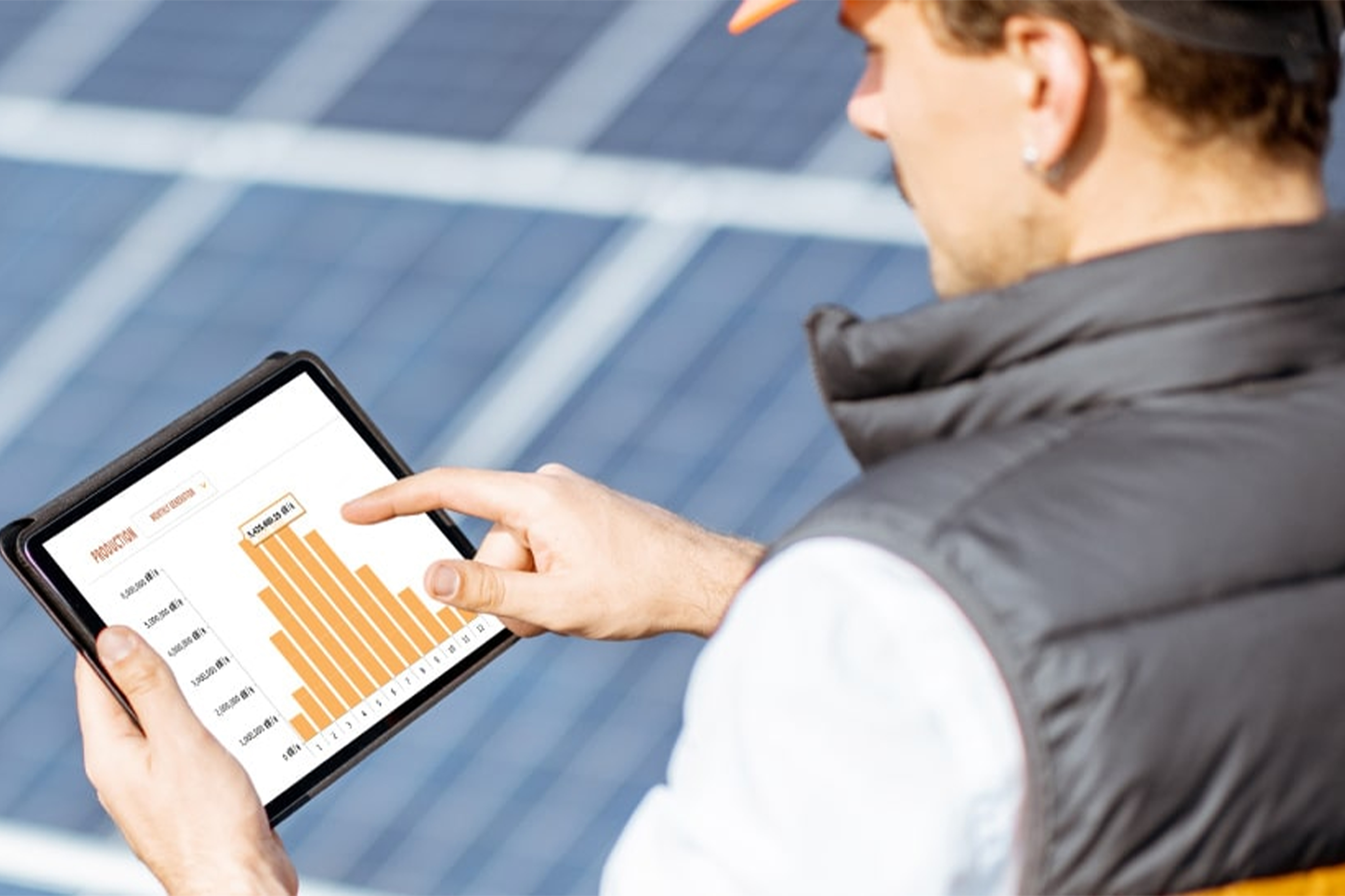 A person stands in front of rows of blue solar panels while holding a tablet. The tablet screen displays an orange bar chart, and the person points at the chart, suggesting monitoring or analyzing solar energy data outdoors.