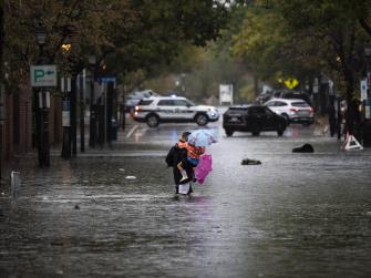 A man carries two children holding colorful umbrellas across a flooded city street.
